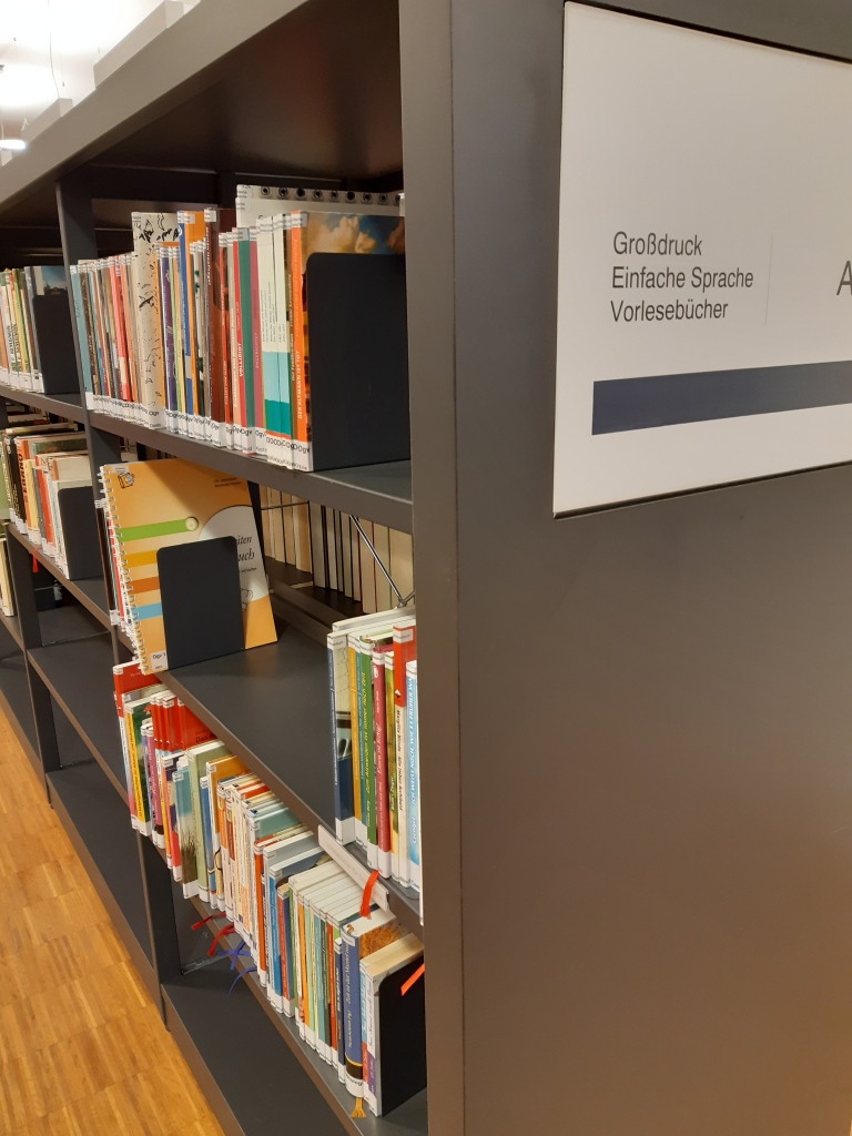 Regal in der Bibliothek, es sind viele schmale Bücher zu erkennen. An der Stirnseite ein Schild "Großdruck, Einfache Sprache, Vorlesebücher"