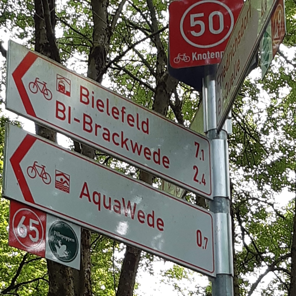 Beschilderung: ganz oben ein rotes Schild mit "Knotenpunkt 50", dann mehrere Richtungsschilder "Bielefeld 7,1" (km), "Brackwede 2,4", "Aquawede 0,7"; darunter 2 quadratische Schilder mit dem Hinweis, dass es nach links zum Knotenpunkt 65 geht und das Logo "Pillepoppen"