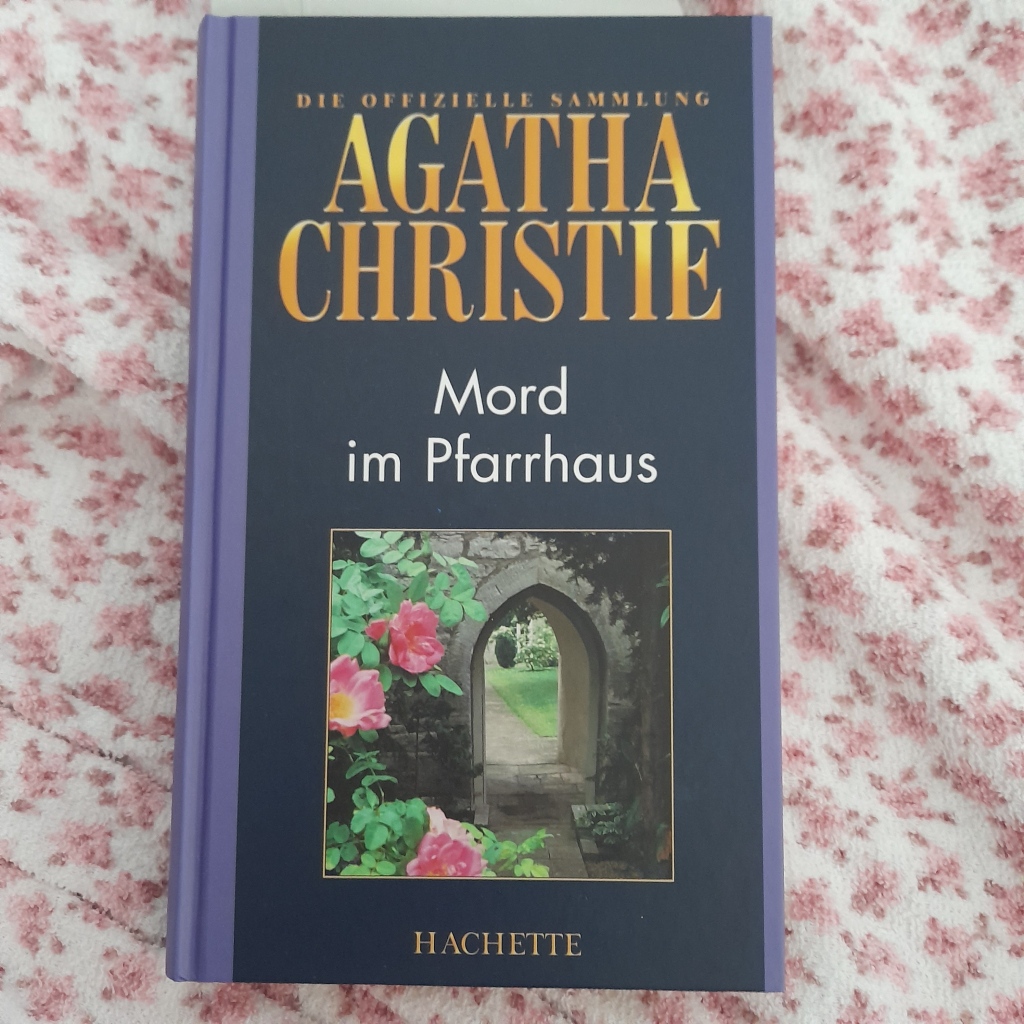 Roman "Mord im Pfarrhaus" von Agatha Chrstie. Das Buch liegt auf einem Frottee-Tuch mit rosa Blütenmuster. Das Titelbild des Romans zeigt ein Steintor mit rankenden Rosen