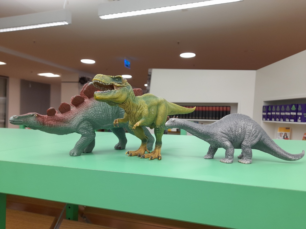 3 Plastik-Dino-Figuren auf einem grünen Brett, Bücherregale im Hintergrund: ein Stegosaurus, ein Tyrannosaurus, ein Brontosaurus (?)