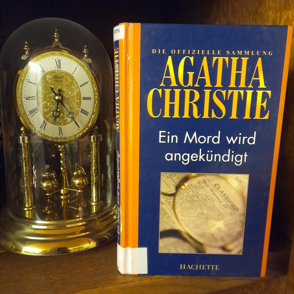 Neben einer alten Tischuhr steht das Buch "Ein Mord wird angekündigt" von Agatha Christie