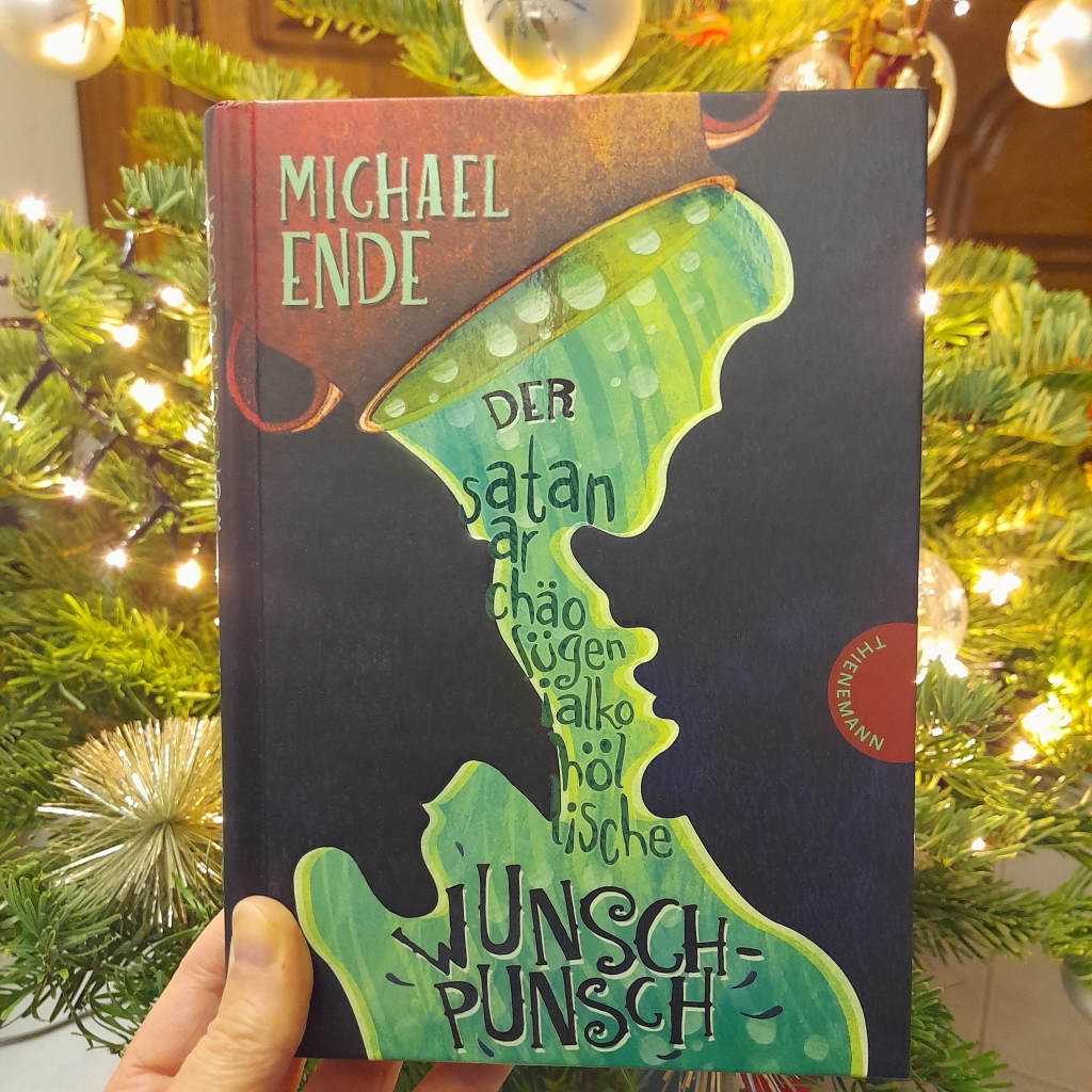 Das Buch "Der satanarchäolügenialkohöllische Wunschpunsch" von Michael Ende wird vor einen beleuchteten Weihnachtsbaum gehalten