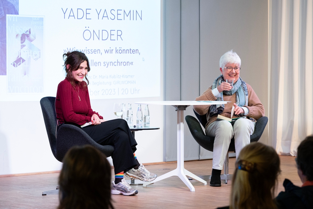 Foto: 2 Frauen, lachend, auf einer Bühne sitzend, schauen ins Publikum. Im Hintergrund der Schriftzug "Yade Yasemin Önder: Wir wissen, wir fallen, und fallen synchron"