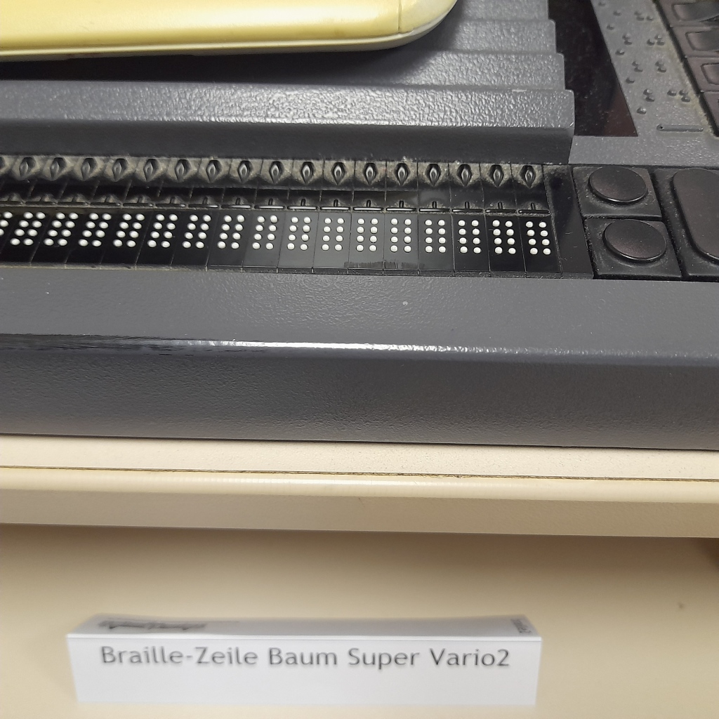 Großaufnahme einer schwarzen Apparatur mit weißen Punkten in Achter-Blöcken nebeneinander (= Stifte, die die Braille-Zeichen bilden können), davor steht ein Schildchen "Braille-Zeile Baum Super Vario2"