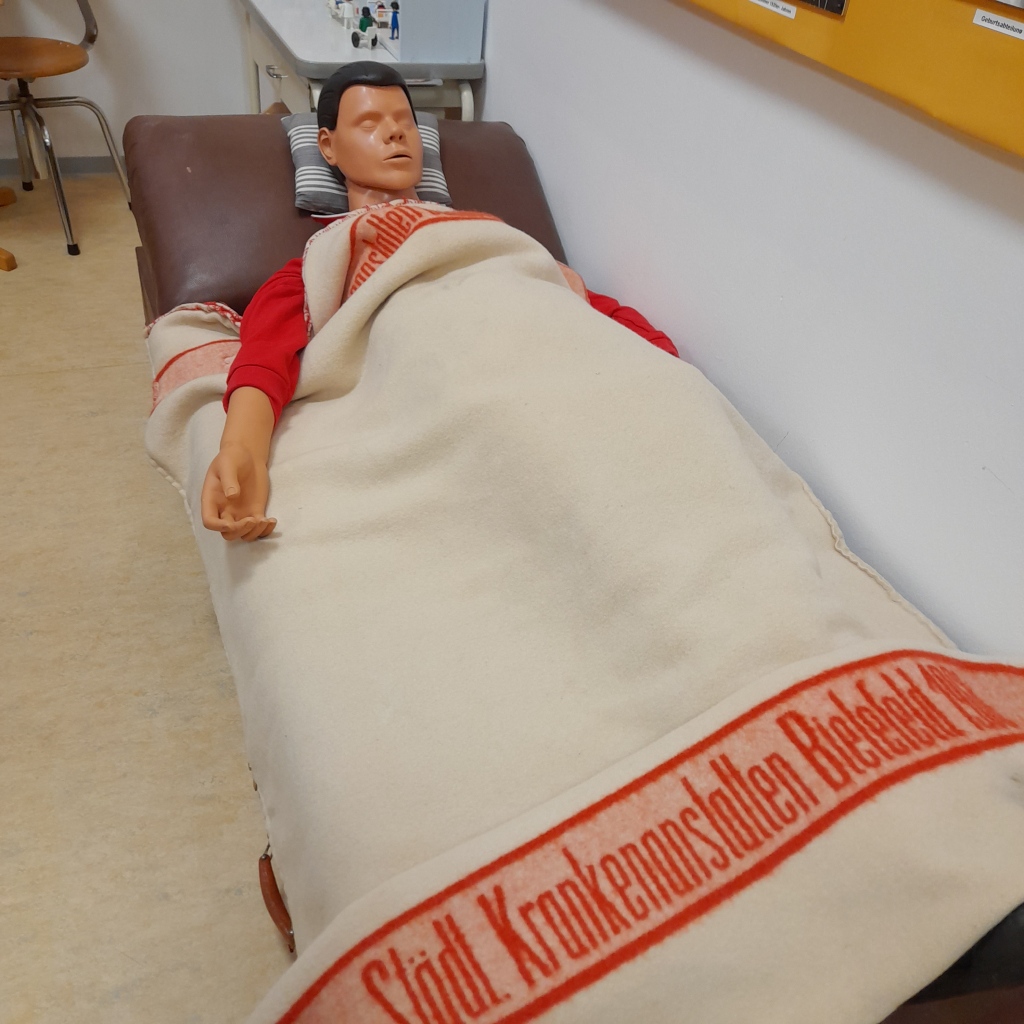 Foto: eine menschliche Puppe liegt auf einer alten Liege, auf der hellen Decke steht "Städt. Krankenanstalten Bielefeld" in roter Farbe aufgedruckt.
