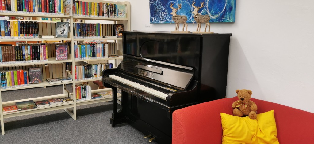 Foto vom Klavier, daneben ein Regal mit Kinderbüchern und auf der anderen Seite ein rotes Sofa mit gelbem Kissen (und dem Teddybär)