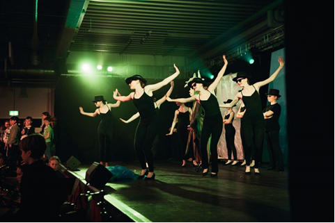 Auf einer Bühne tanzt eine Gruppe Frauen mit schwarzen Trikots, Hosen und Zylinder, vor der Bühne Publikum